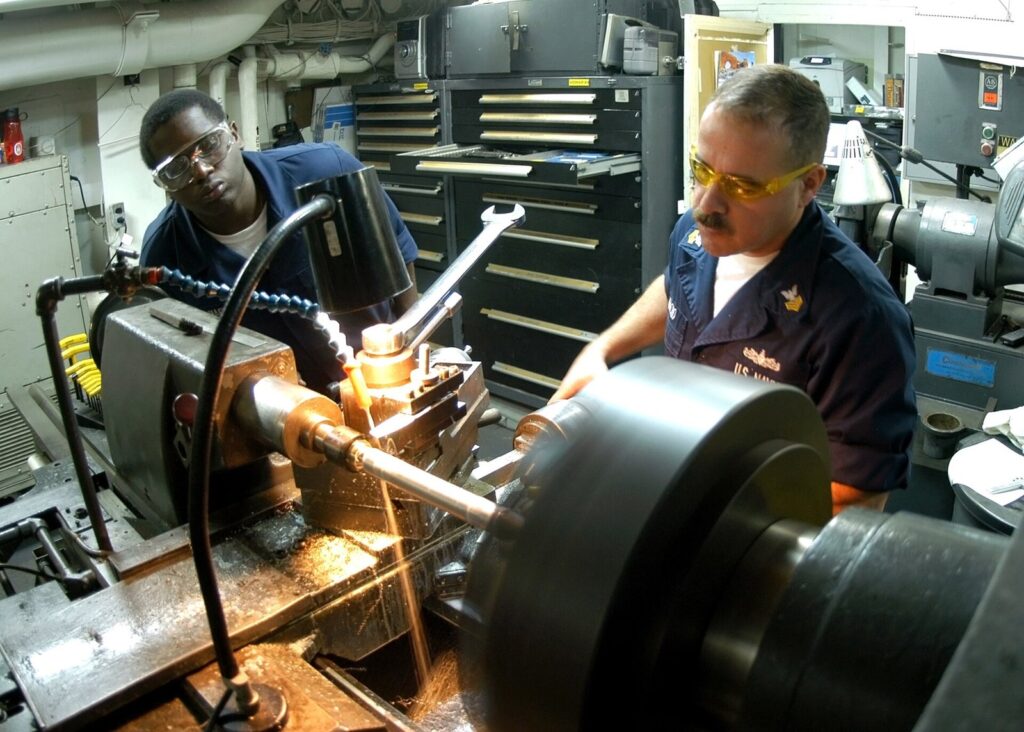 Two machinists cutting a shaft on a lathe in a ship's machine shop