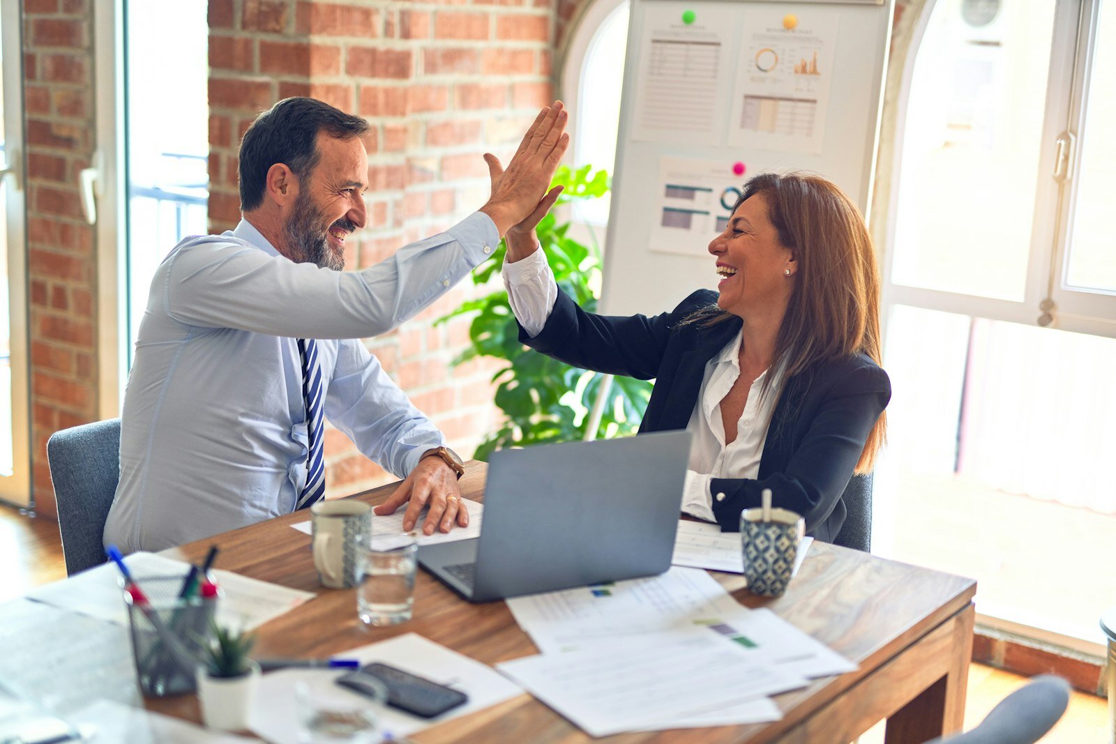 Two colleagues high-fiving at a desk with whiteboard diagrams—enterprise collaboration on process decisions. Photo: Unsplash.