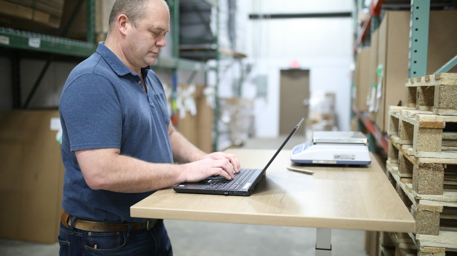 Warehouse associate working at a standing desk with a laptop beside a shipping scale—the frontline service and logistics moment industrial CRM agents must reason through. Photo: Unsplash.