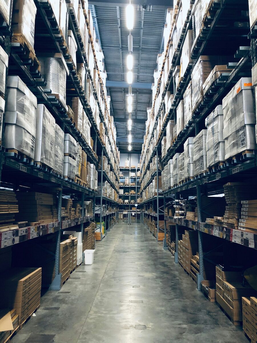 Long aisle of industrial warehouse racking packed with palletized inventory under overhead lights. Photo: Unsplash.
