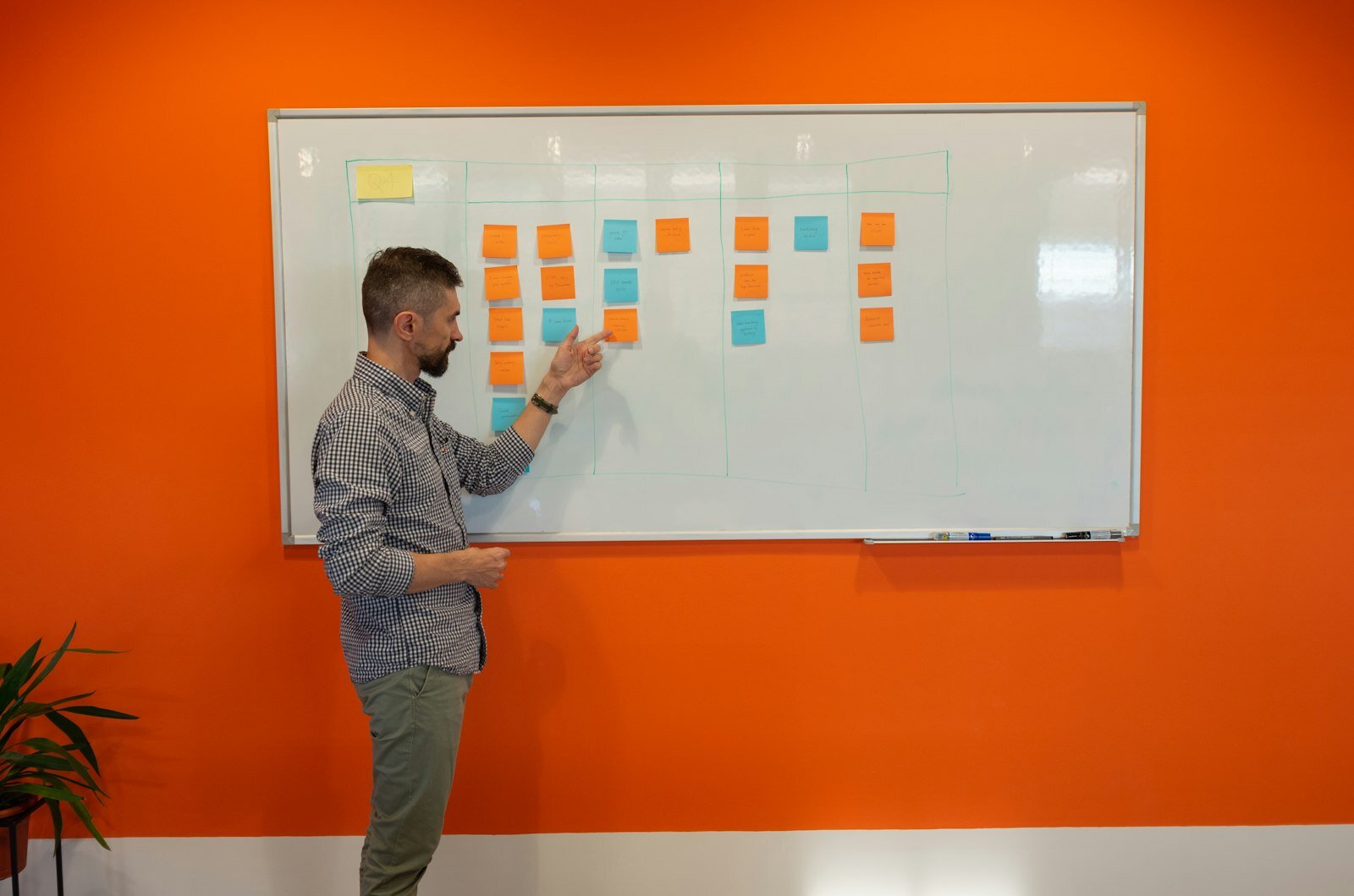 Program lead pointing at a sticky-note planning board in a meeting room—portfolio intake still dies without one system of record. Photo: Paymo / Unsplash.