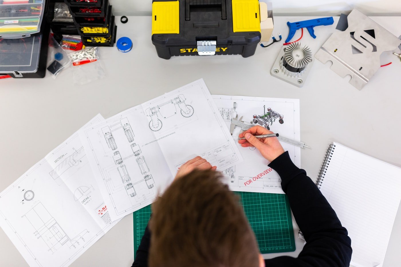 Engineer reviewing robot vehicle blueprints and calipers on a workbench with parts bins. Photo: Unsplash.