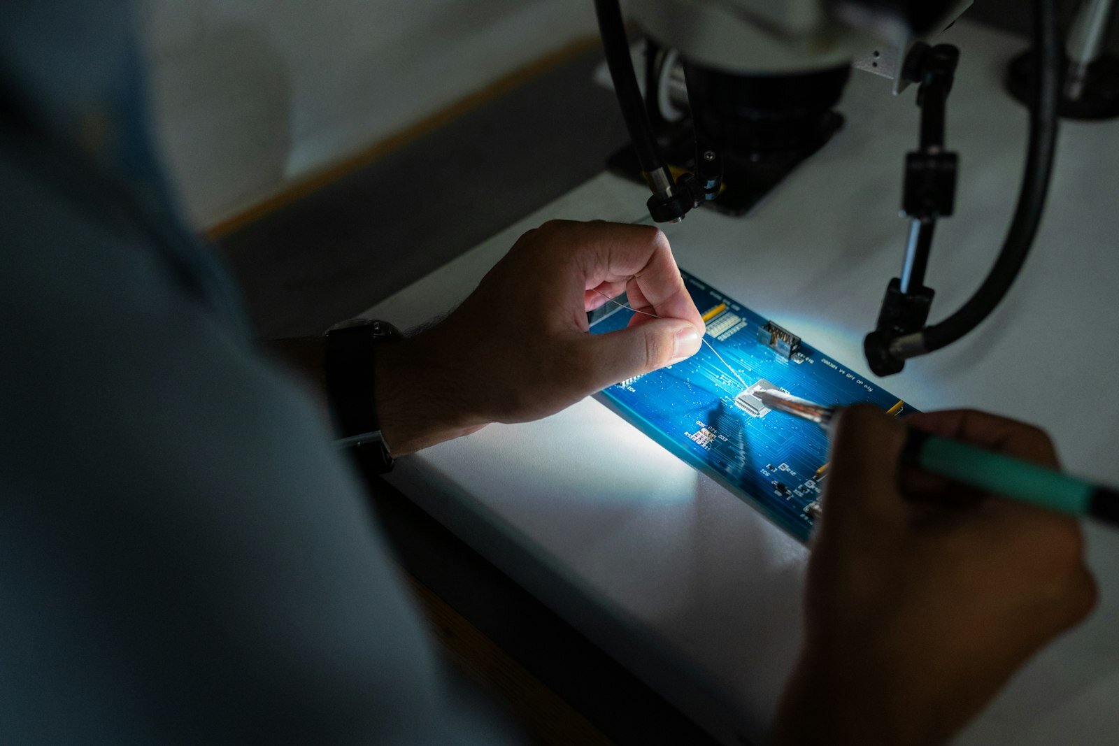Technician soldering a circuit board under a work lamp—precision repair that digital procedures and required checks are meant to standardize. Photo: Unsplash.