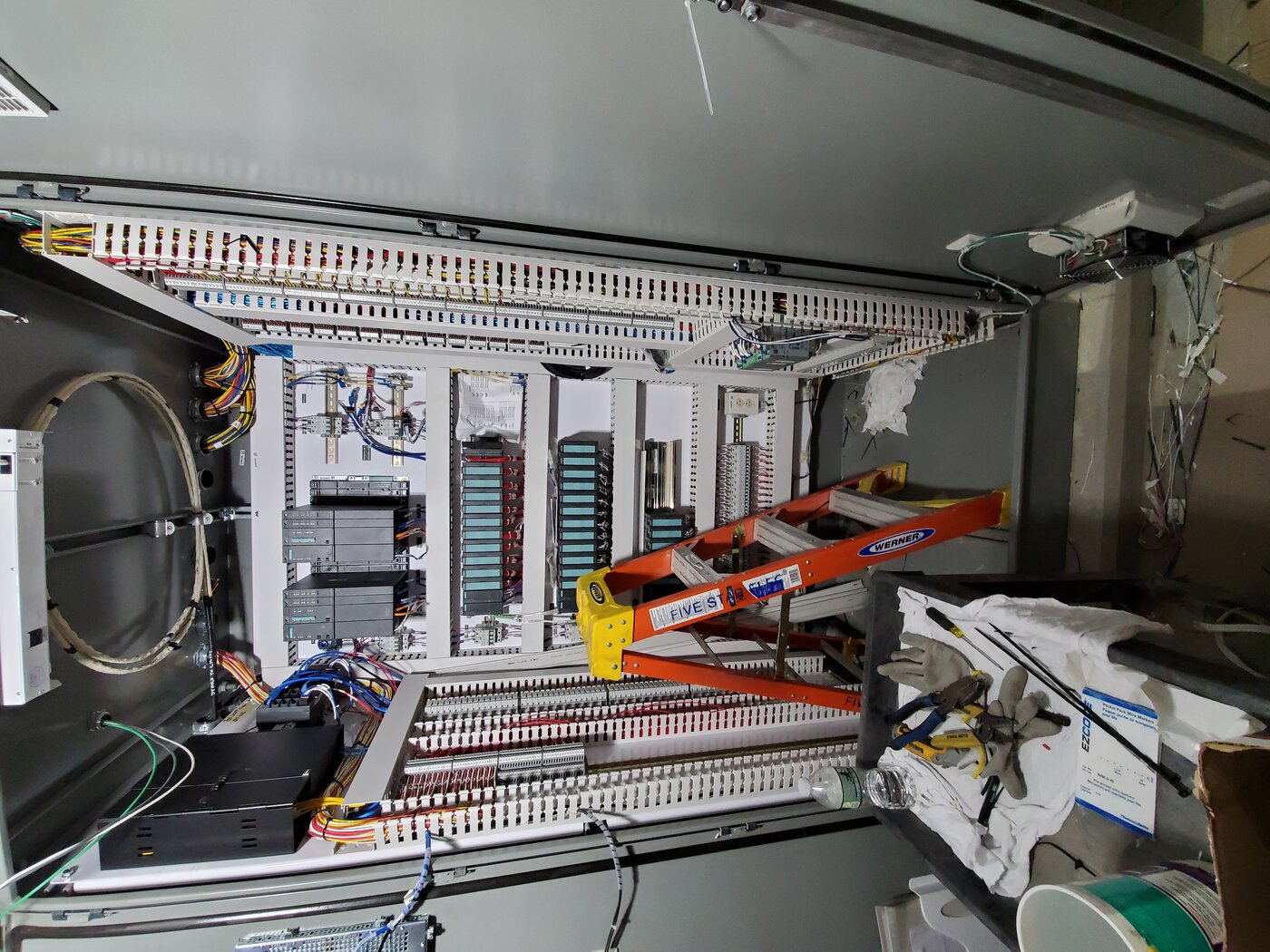 Open PLC control panel with industrial wiring, DIN-rail modules, and computer control systems in a mechanical room. Photo: Wikimedia Commons.