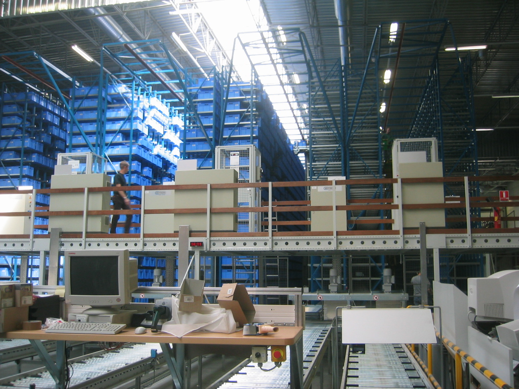 Rows of blue plastic bins stacked high in an automated small-parts warehouse, with workstations on a mezzanine in front.