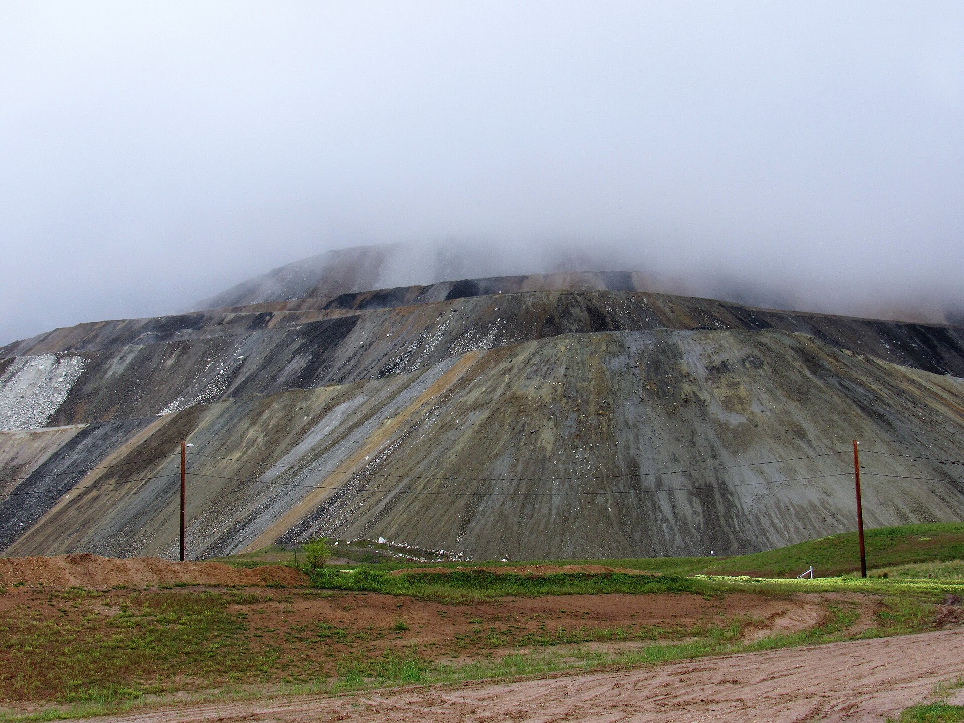 The terraced gray and tan waste-rock slopes of the Bingham Canyon copper mine in Utah, with low cloud over the top and green grass in the foreground.