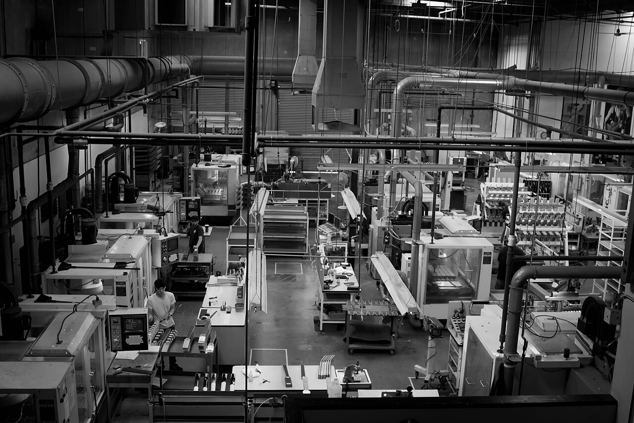 A black-and-white view of a factory floor lined with enclosed CNC milling machines and overhead ducting.