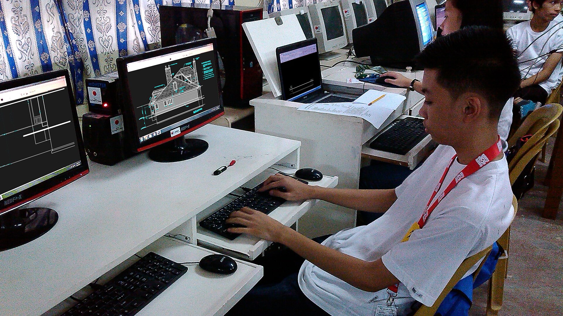 A student in a white shirt with a red lanyard works at a desktop computer in a school computer lab, with CAD drawings displayed on several monitors.