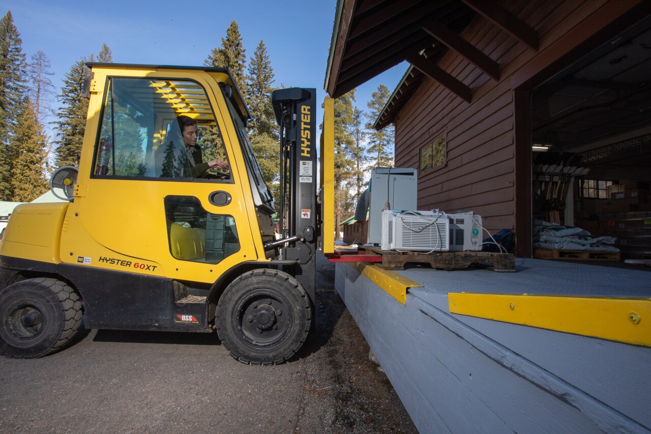 A supply technician drives a yellow forklift carrying pallets of supplies up to a loading dock at a wooden building.