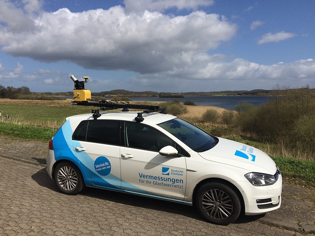A white mobile mapping car with a roof-mounted laser scanner parked on a country road.