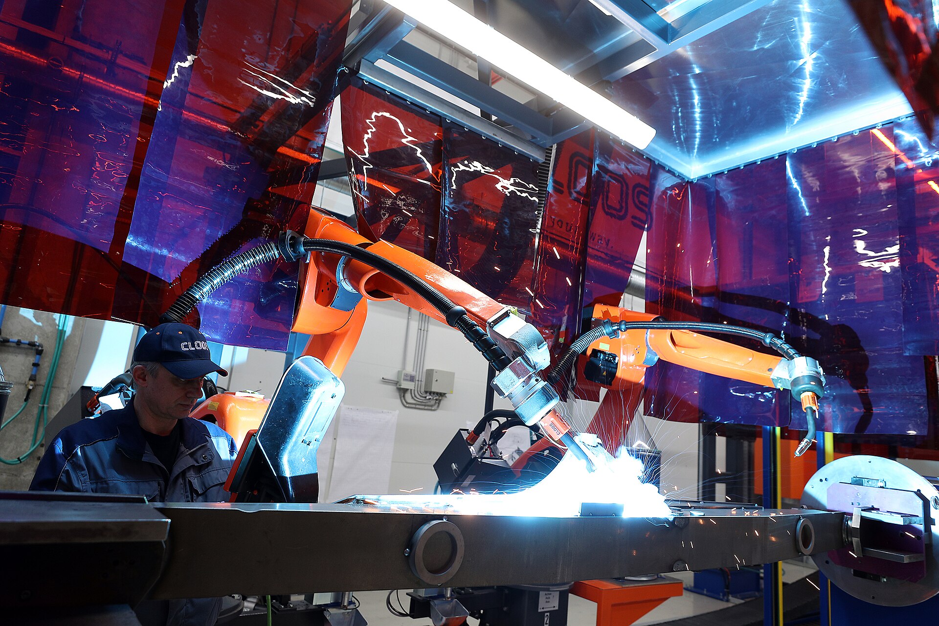 A worker watches two orange robotic arms weld a long steel beam inside a welding cell.