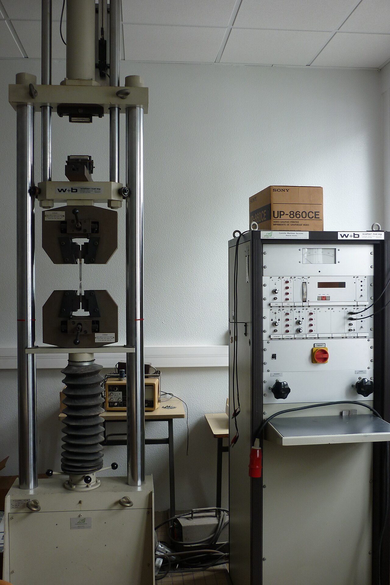 Universal tensile testing machine with a metal specimen clamped in its grips beside its control cabinet