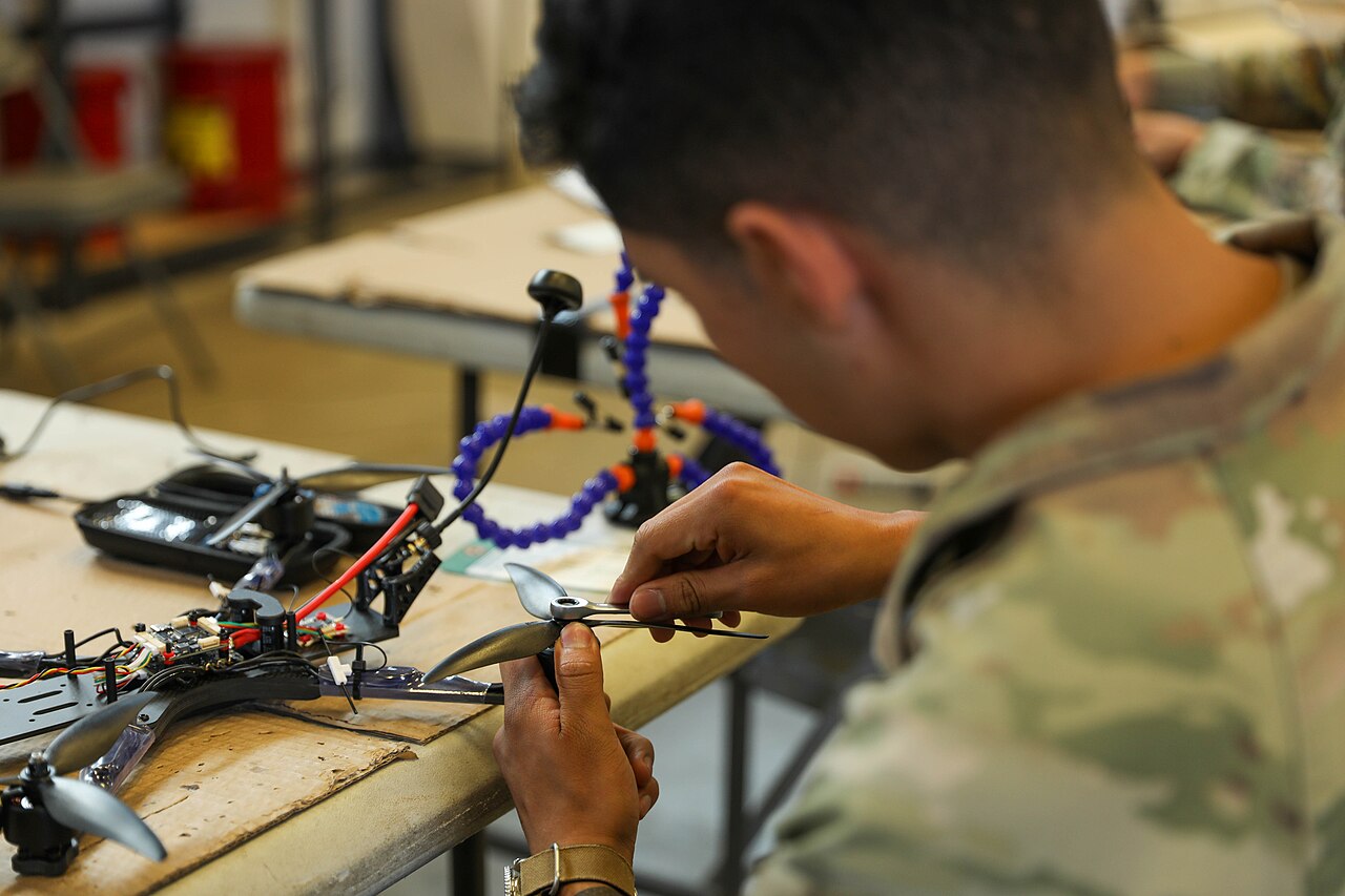 A U.S. Army soldier assembles a small drone at a workbench covered with parts and tools.