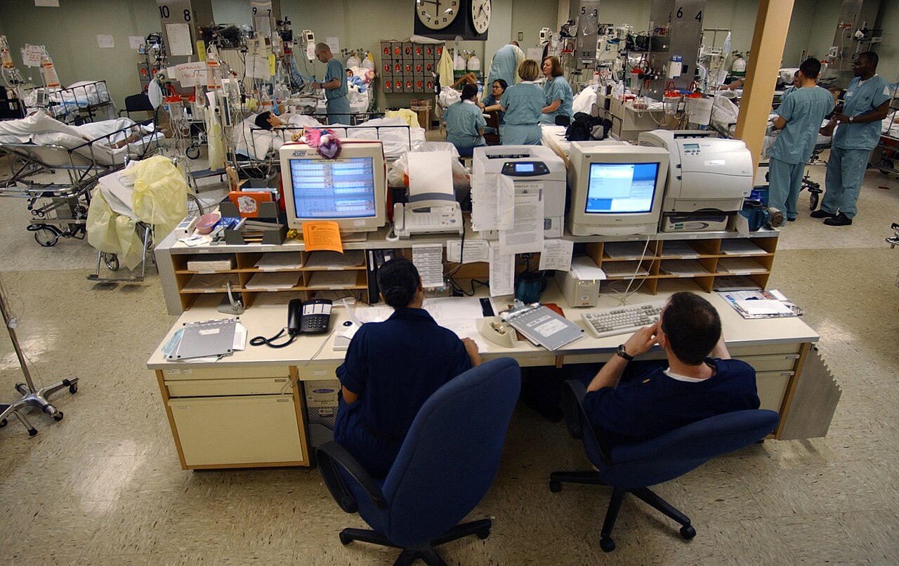 Two staff members sit at a central desk with several computer monitors showing patient heart rates, with hospital beds and nurses in the ward behind them.