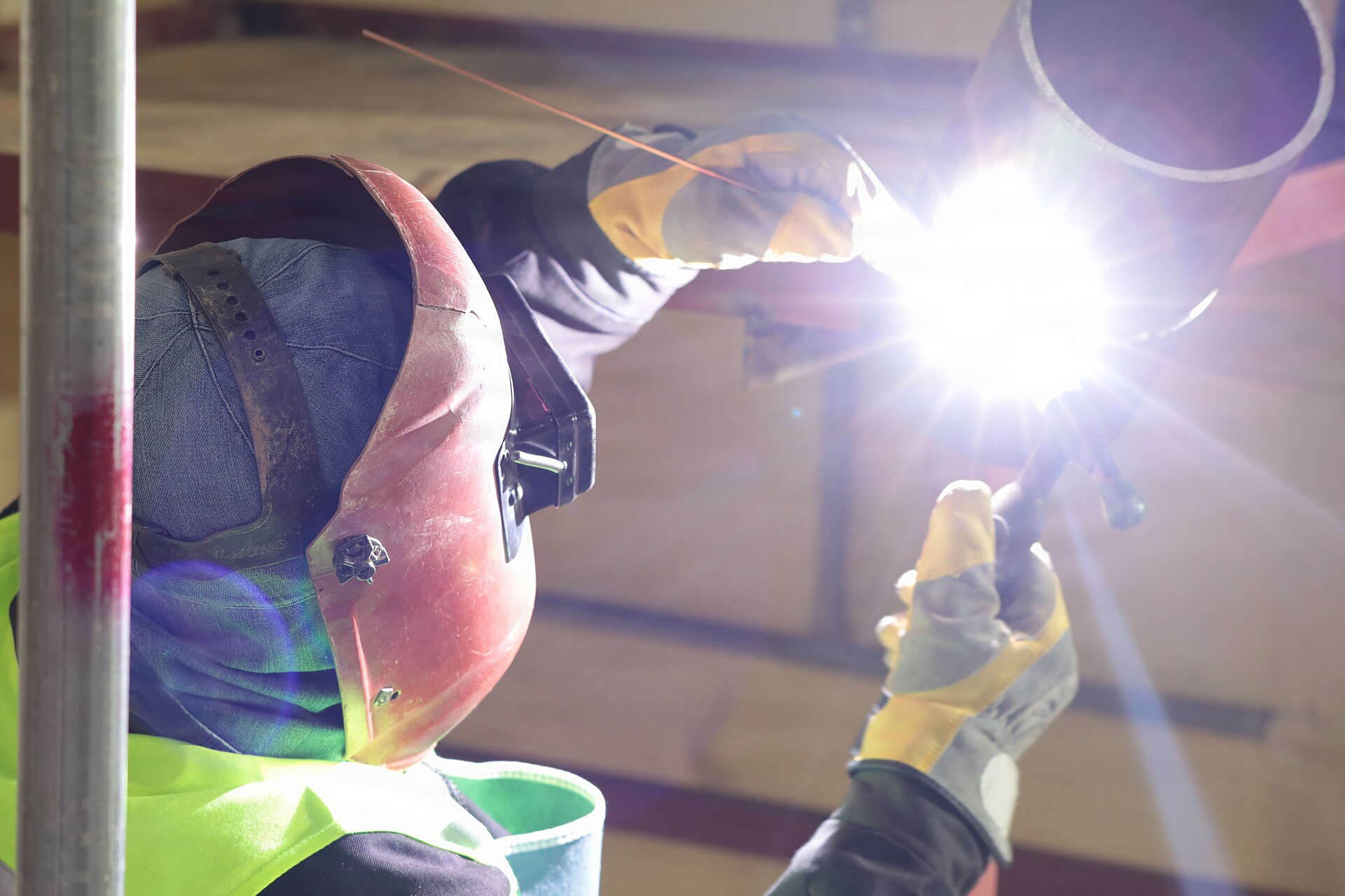 A welder in a hood and gloves runs a gas tungsten arc weld on a thick pipe during a welder qualification test.