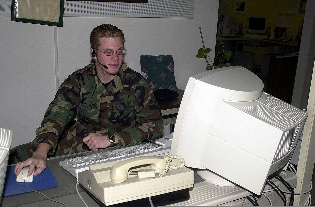 A man in a camouflage uniform wearing a telephone headset works at a desktop computer next to a desk phone.