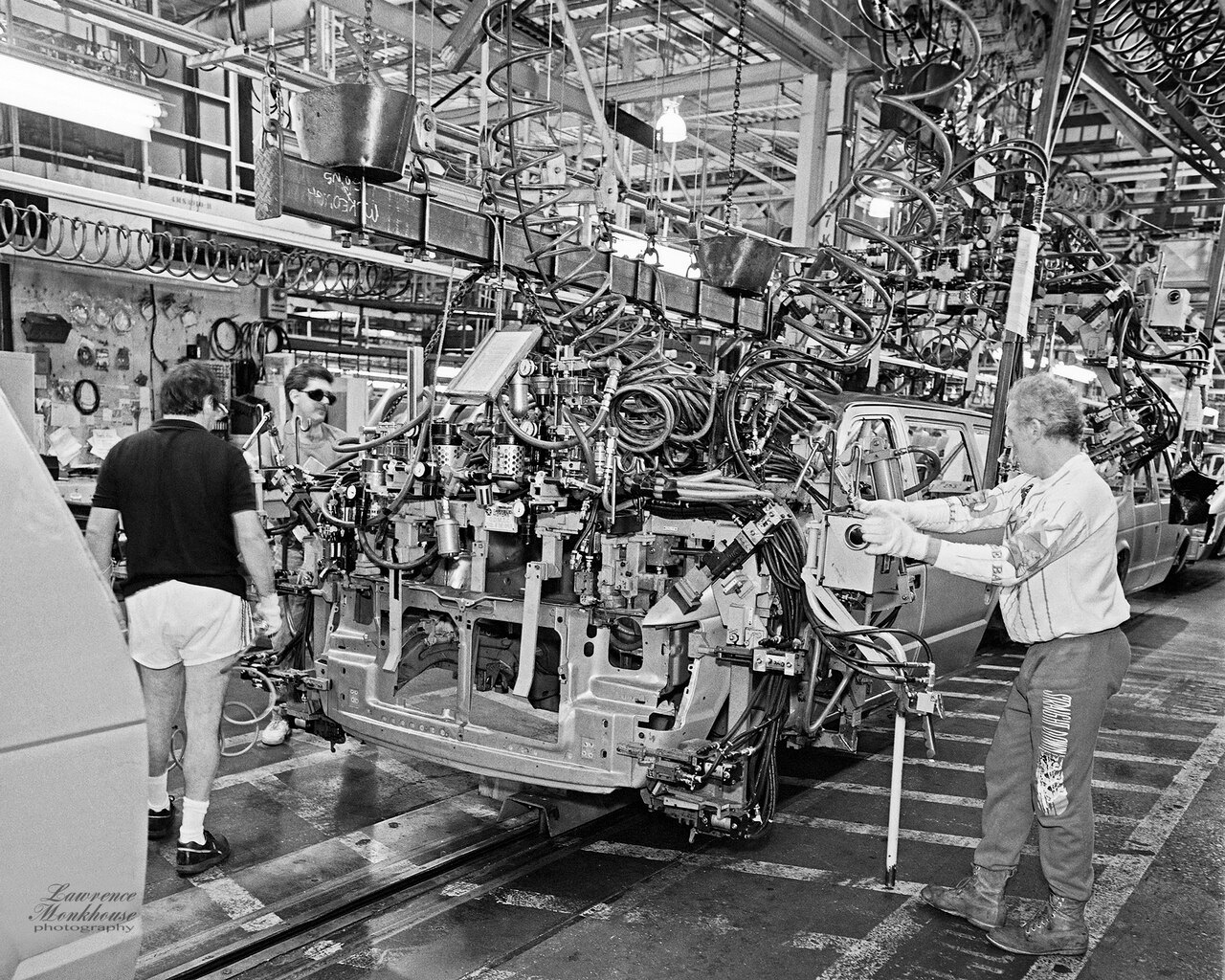 Black-and-white photo of workers fitting parts to an unpainted minivan body shell on an assembly line, surrounded by hoses and tooling.