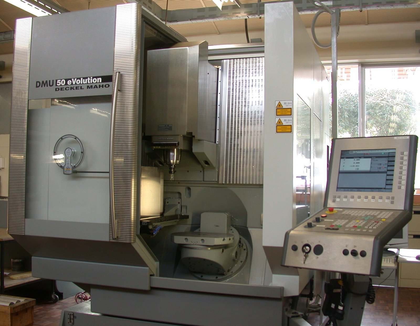 A Deckel Maho DMU 50 eVolution five-axis machining center with its door open, showing the spindle above a rotary table, beside its CNC control panel. Photo: Glenn McKechnie, CC BY-SA 2.0, via Wikimedia Commons.