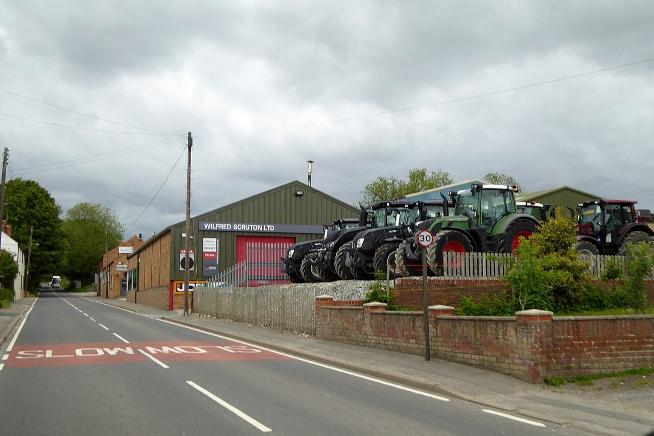 A row of green and red tractors parked in front of a farm equipment dealership on a rural English road.