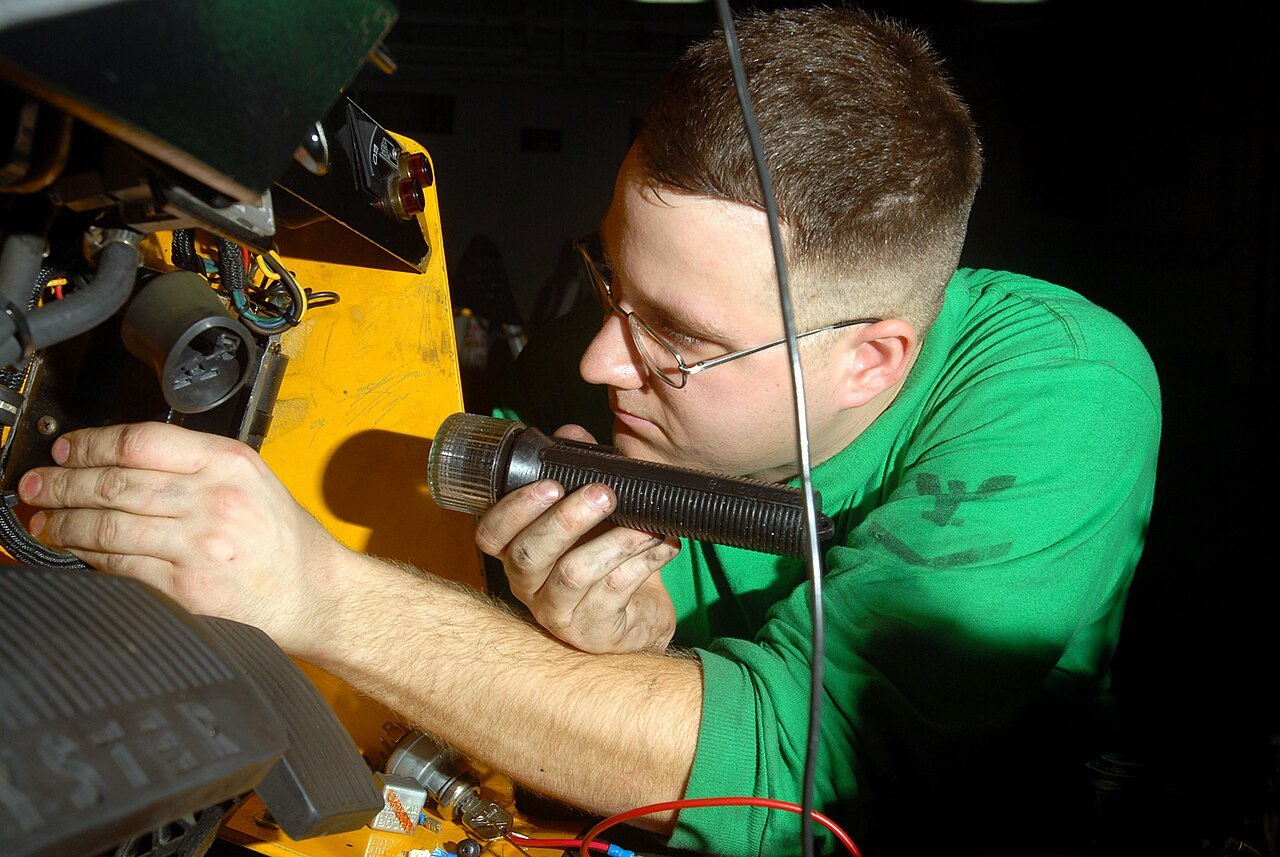 A sailor in a green shirt shines a flashlight into a forklift engine compartment while checking an ignition switch.
