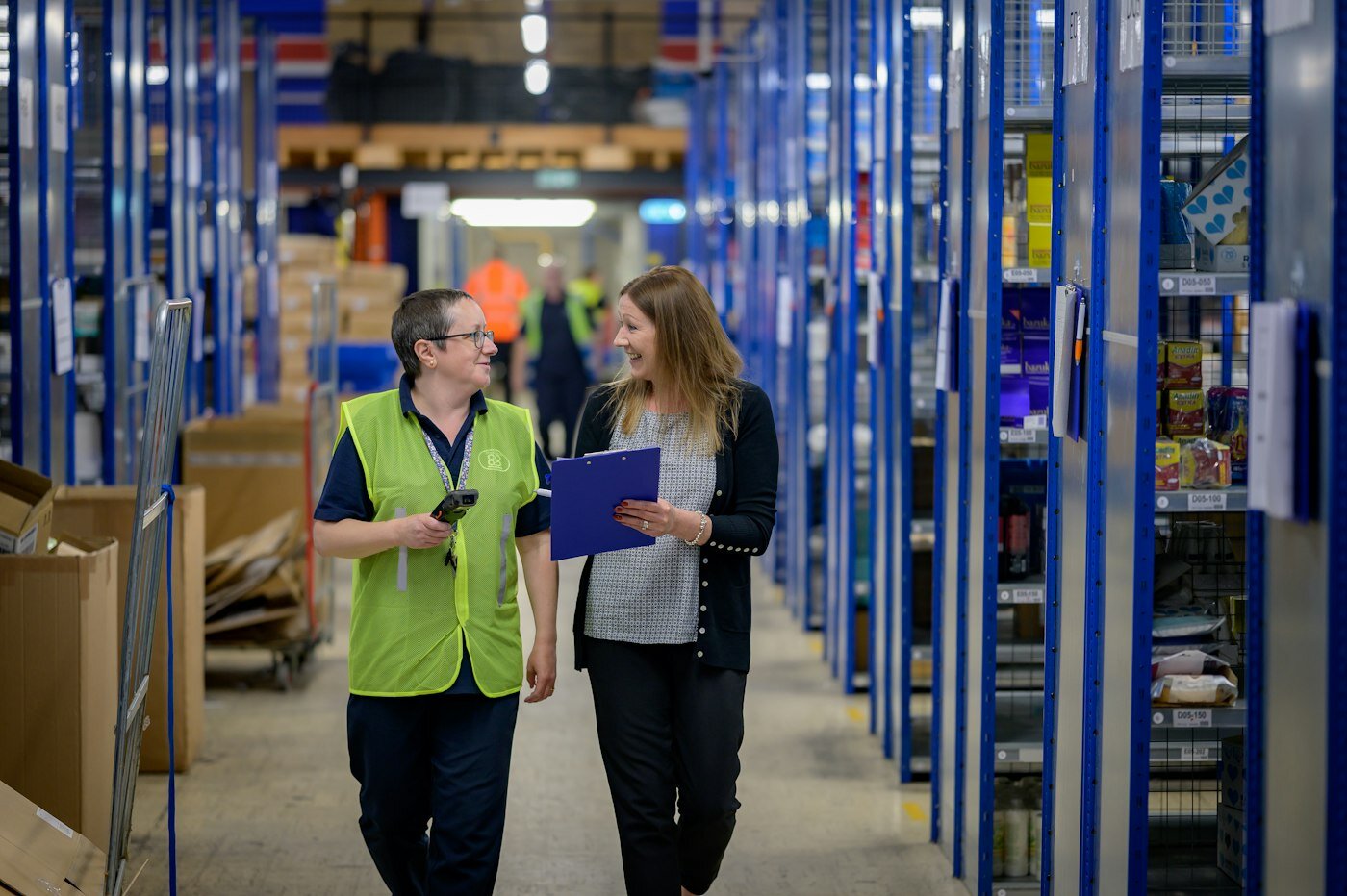 Two people walking a warehouse aisle with a clipboard—where ATP, picking, and production material decisions meet inventory. Photo: Centre for Ageing Better / Unsplash.