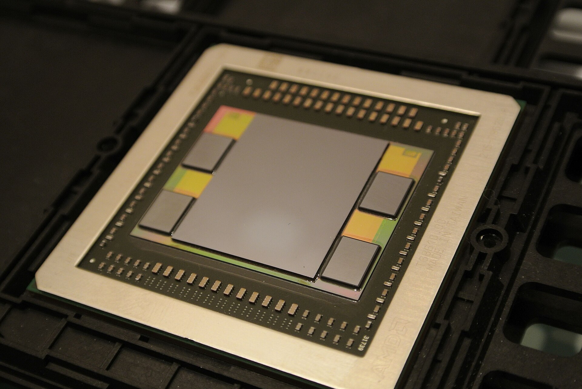 An AMD Fiji GPU package in a carrier tray, showing the large GPU die flanked by four HBM memory stacks on a silicon interposer, set on the package substrate with a metal stiffener ring. Photo: C. Spille/pcgameshardware.de, CC BY-SA 4.0, via Wikimedia Commons.