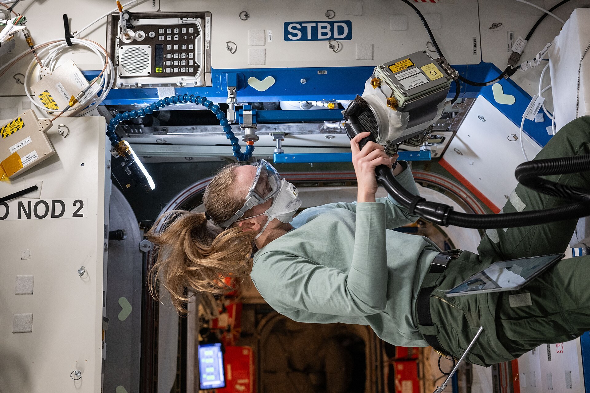 NASA astronaut Nichole Ayers, wearing goggles and a face mask, holds a vacuum hose to a ventilation fan assembly inside the International Space Station's Destiny laboratory module. Photo: NASA, public domain, via Wikimedia Commons.
