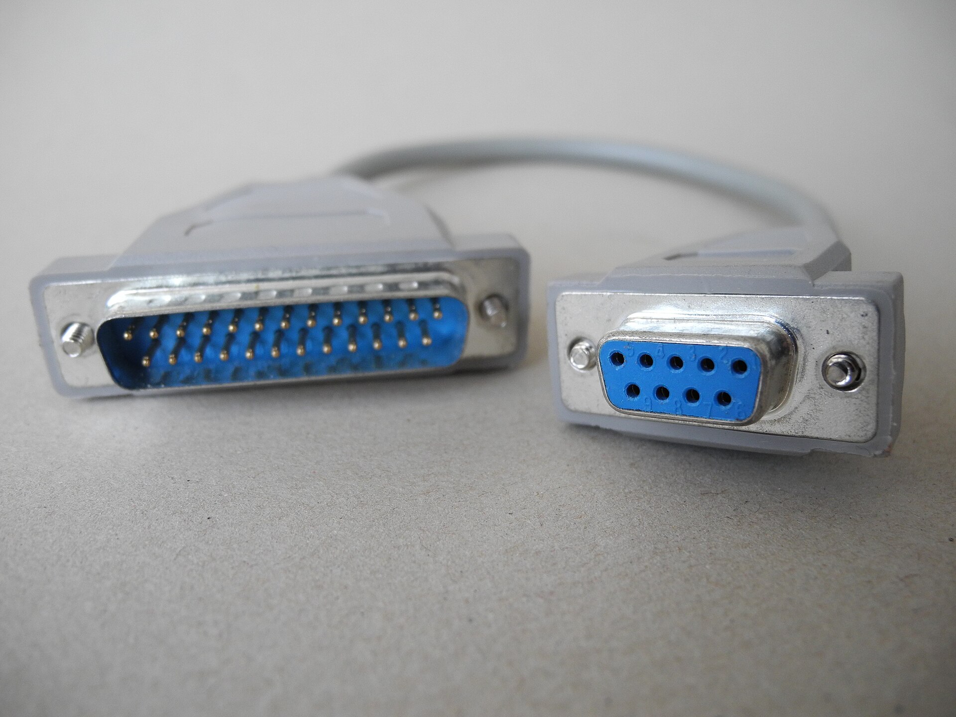 A gray RS-232 serial adapter cable with a 25-pin male D-sub connector on one end and a 9-pin female D-sub connector on the other. Photo: Hubert Berberich (HubiB), public domain, via Wikimedia Commons.