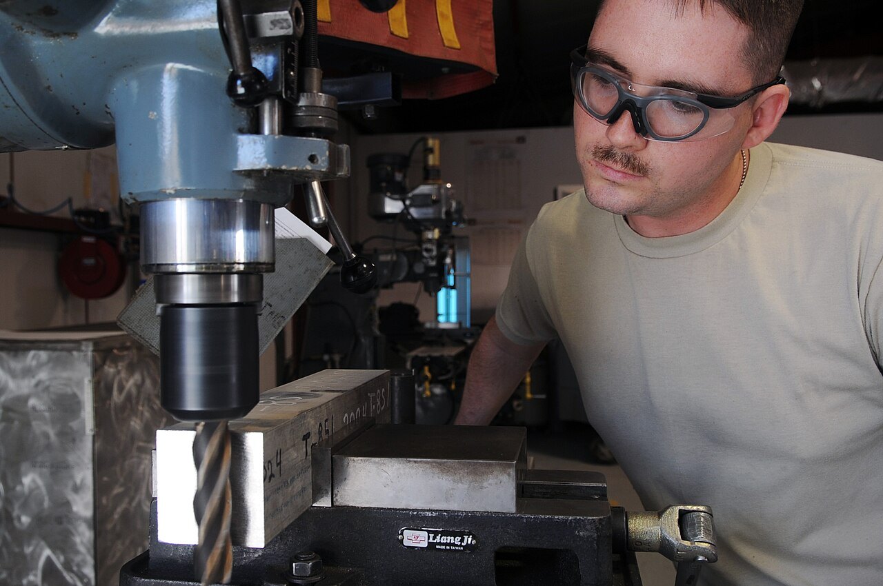 A metal technician wearing safety glasses faces down an aluminum block on a milling machine.