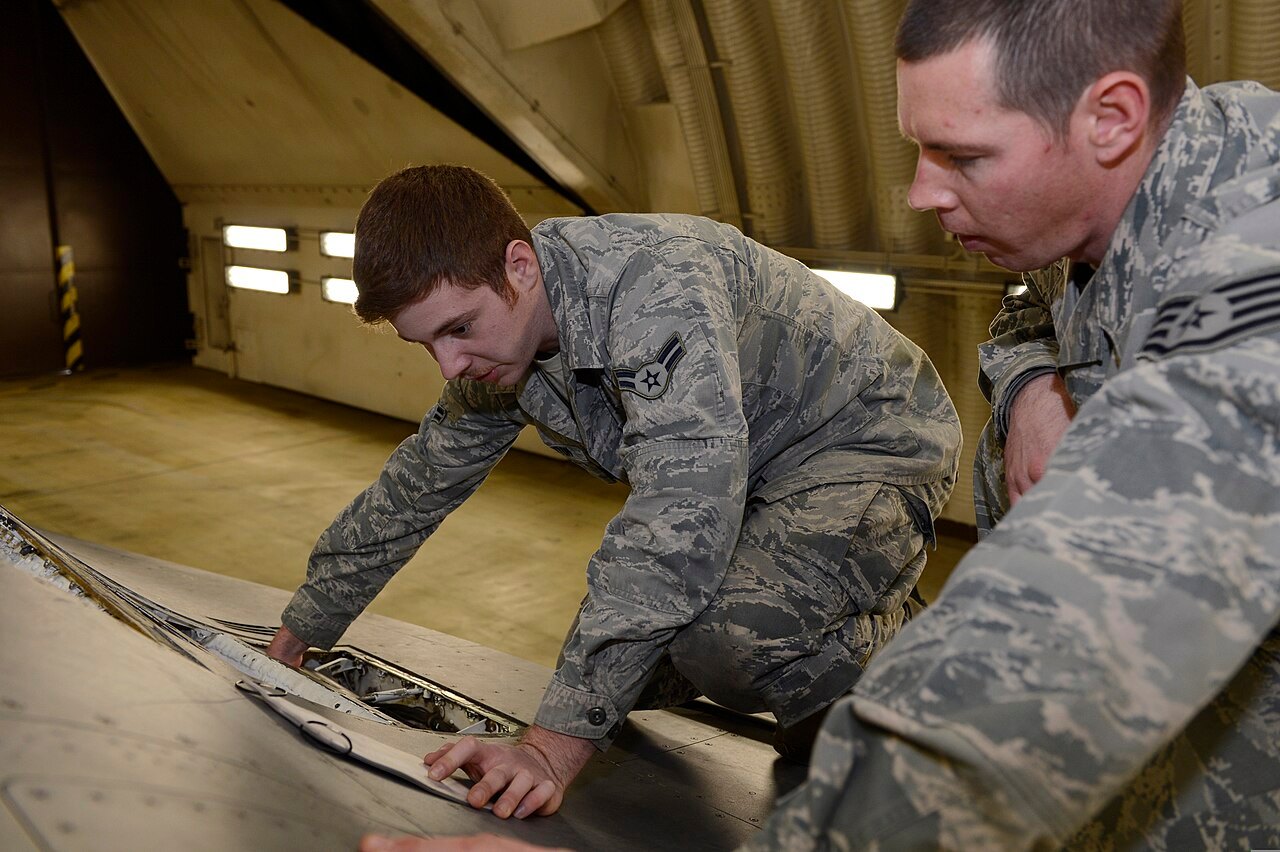 Two U.S. Air Force weapons technicians kneel under an F-16 and read a printed checklist while inspecting its weapons system.
