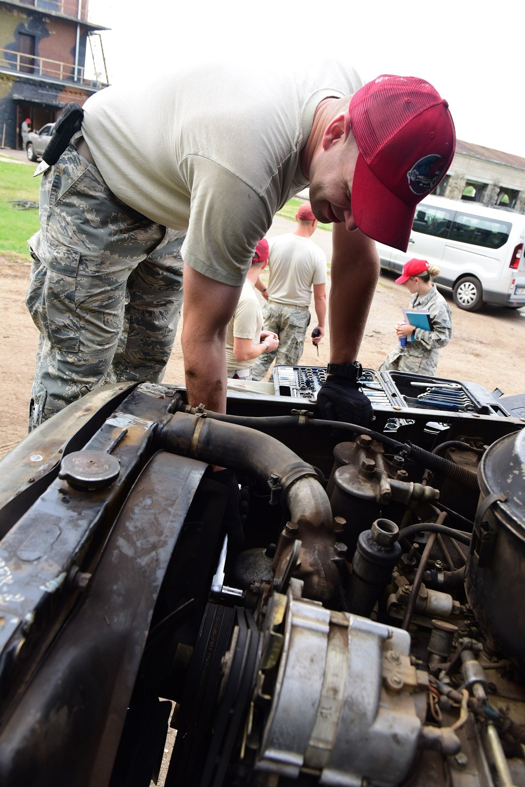 An Air National Guard vehicle maintenance technician leans over the open engine bay of a water truck during a preventive maintenance inspection, with other airmen and a clipboard in the background. Photo: U.S. Air National Guard / Tech. Sgt. Claire Behney, public domain, via Wikimedia Commons.